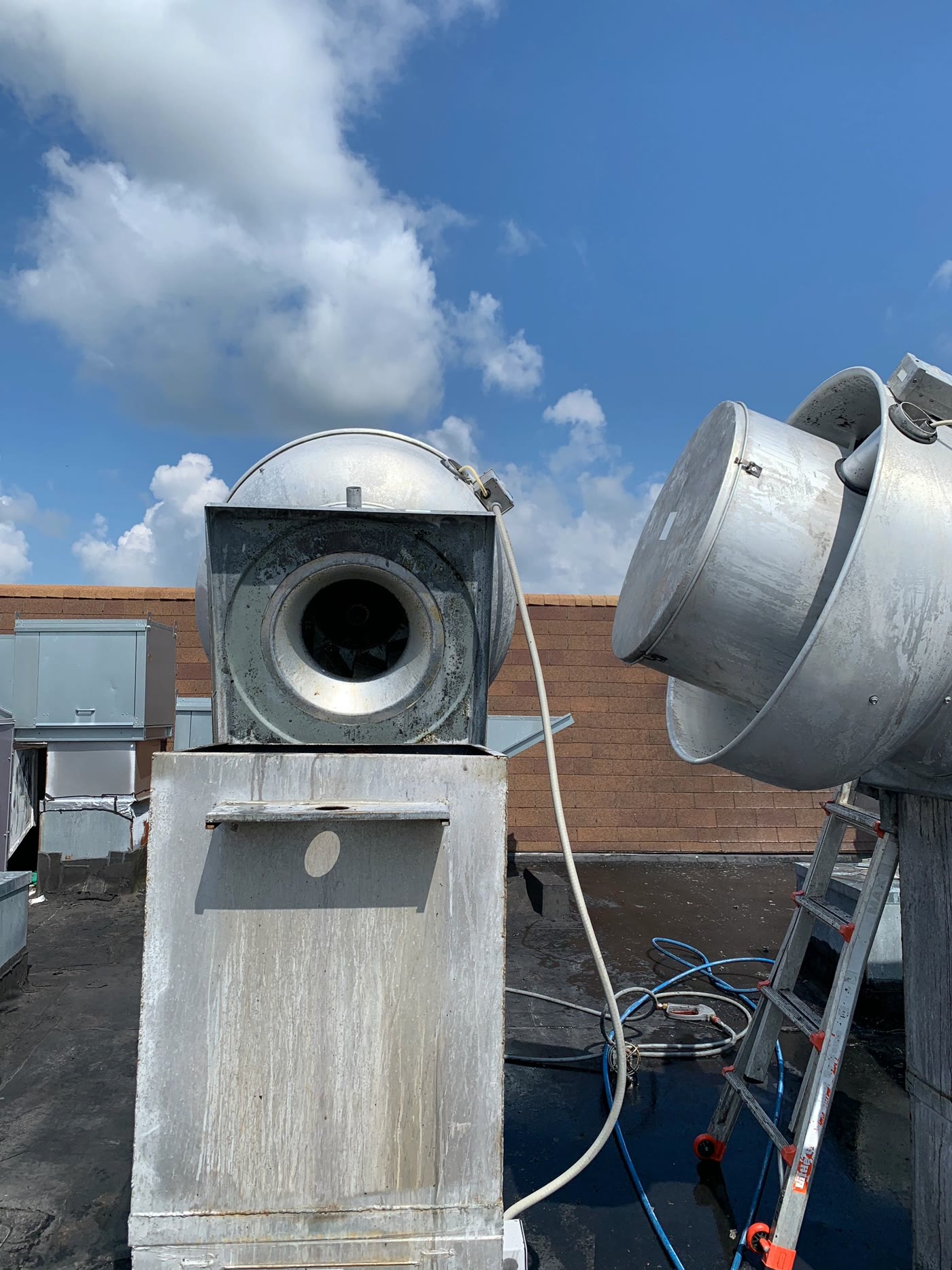 A grease-stained rooftop exhaust fan tipped open on its hinge against a cloudy sky
